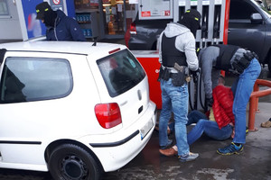 Policjanci przeszukują zatrzymanego mężczyznę siedzącego na ziemi na terenie stacji benzynowej.