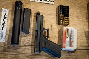 Pistolet, magazynek, tłumik, pudełko po amunicji, plastikowy koszyczek wypełniony amunicją oraz skalówki leżące na blacie stołu.