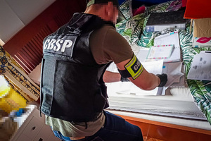 Policjant w kamizelce i opasce z napisem &quot;CBŚP&quot; przegląda dokumentację leżącą na łóżku.