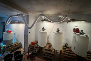 Cztery plastikowe beczki wypełnione cieczą stojące w pomieszczeniu. Nad nimi pod sufitem zamontowana prowizoryczna instalacja wentylacyjna.