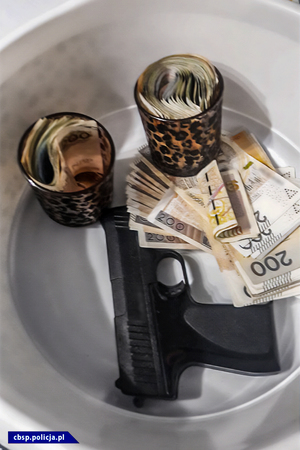 Szklanki wypełnione banknotami, obok leżące banknoty o nominale 200 PLN oraz pistolet.