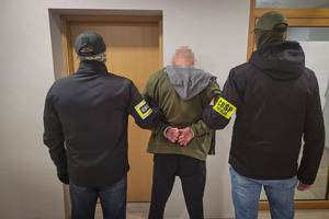 Policjanci z opaskami na ramionach z napisem CBŚP, trzymają pod ręce zatrzymanego.