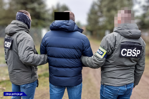 Funkcjonariusze w kurtkach z napisem CBŚP, trzymają pod ręce zatrzymanego.