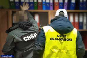 Funkcjonariusze CBŚP i Służby Celno-Skarbowej przeszukujący regał z segregatorami.