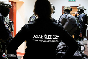 Kobieta w kurtce z napisem &quot;Dział śledczy - Polska Agencja Antydopingowa&quot;, w tle umundurowani funkcjonariusze CBŚP.